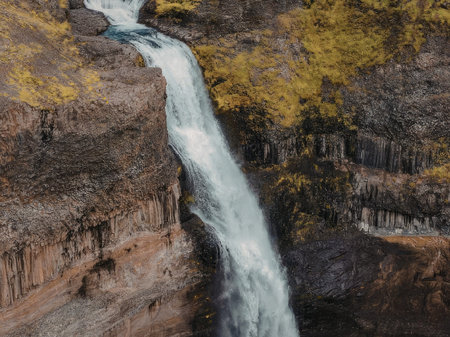 A serene waterfall flows down a rugged cliffside, surrounded by lush greenery and rocky terrain,...