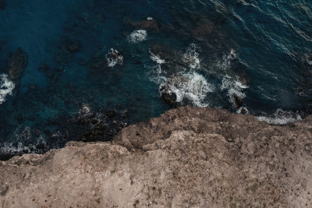 Aerial view of ocean waves crashing on a rocky shoreline, showcasing the powerful blue water and...