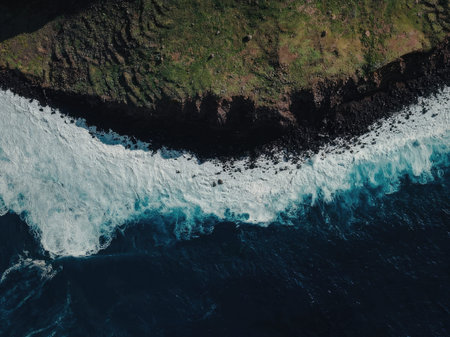 Aerial view of a wave crashing against a cliffside with lush greenery on top