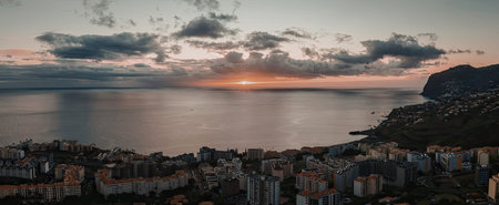A serene coastal cityscape with buildings and mountains under a vibrant orange and blue sky at...