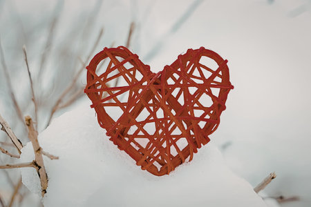A red heart-shaped decoration made of woven material is placed on a snowy surface, surrounded by...