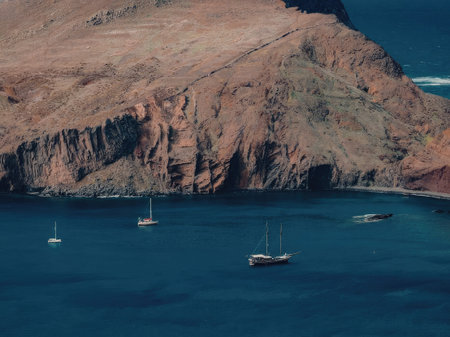 The image depicts sailboats on a calm blue body of water with a large cliffside in the background. The cliffside is brown and green with a few patches of grass. The water is a deep blue color and appears to be very calm.の写真素材