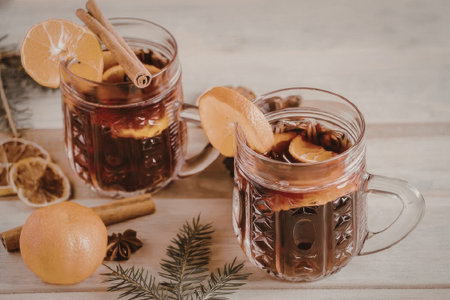 Two glass mugs of mulled wine or punch with orange slices and cinnamon sticks on a wooden table with...