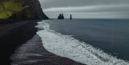 A wave crashes onto a black sand beach with a cliff and rock formations in the background.