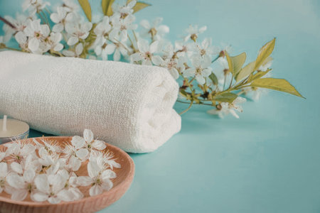 A serene spa setup featuring a rolled white towel, a wooden bowl with flowers, and a sprig of...