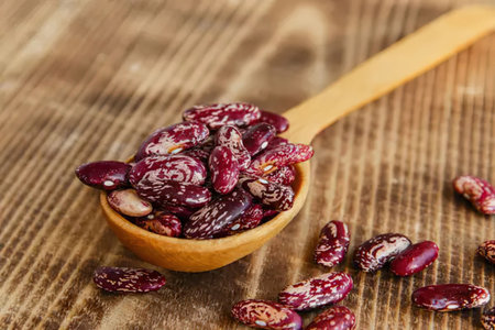 A wooden spoon filled with dried red beans on a wooden surface with some scattered around it.