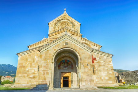 A historic stone church with a large archway and a cross on top, set against a clear blue sky and...