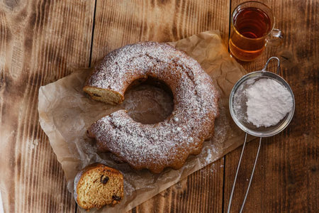A bundt cake with powdered sugar, a cup of tea, and a muffin on a wooden table.