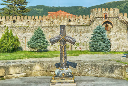 A large Celtic cross stands in front of a historic stone castle with a mountainous backdrop.