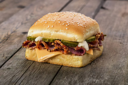 A chicken sandwich with cheese, bacon, and pickles on a sesame seed bun, placed on a rustic wooden table.の写真素材