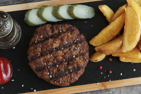 A grilled burger patty served with fries, cucumber slices, and condiments on a black tray.