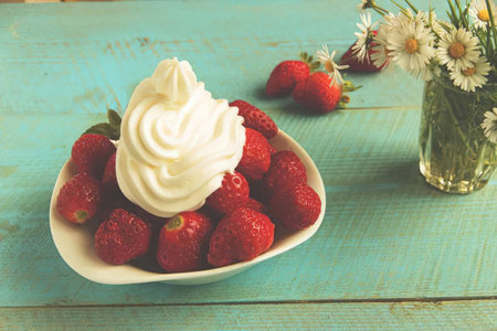 A plate of fresh strawberries topped with whipped cream on a rustic blue wooden table with a vase of...