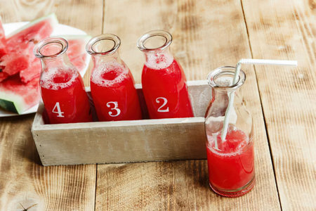 A wooden tray holding three glass bottles filled with red watermelon juice, each labeled with a number, and a single bottle with a straw beside it.の写真素材