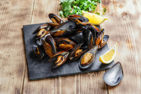 A plate of freshly cooked mussels served on a black slate with lemon wedges and herbs on a wooden...