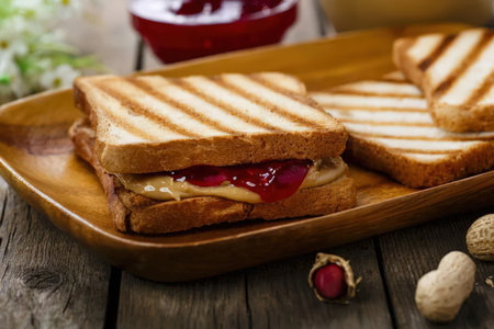 A grilled peanut butter and jelly sandwich cut in half on a wooden plate with peanuts and a bowl of jelly in the background.の写真素材