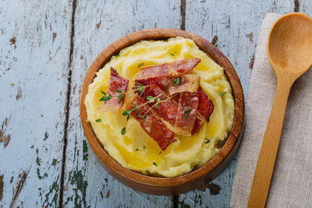 A bowl of mashed potatoes topped with crispy bacon and fresh herbs, served on a rustic wooden table with a wooden spoon.の写真素材