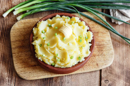 A bowl of mashed potatoes with butter and chives on a wooden cutting board