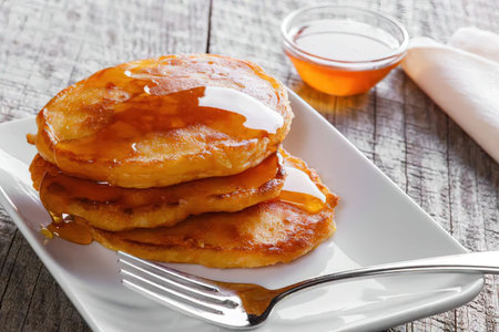 A stack of pancakes on a white plate with syrup drizzled on top, accompanied by a fork and a small...