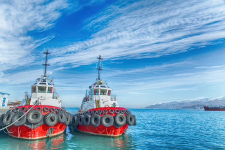 Two red and white tugboats are moored in calm waters, with a mountain range in the background under a blue sky with wispy clouds.の写真素材