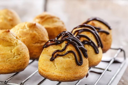 A close-up view of chocolate profiteroles on a cooling rack, with some topped with chocolate...