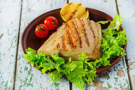 A grilled chicken breast on a brown plate with lettuce, tomatoes, and grilled pineapple on a white wooden table.の写真素材