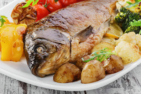 A plate of grilled fish served with roasted potatoes, tomatoes, and broccoli on a rustic wooden table.の写真素材