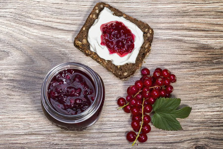 A jar of red currant jam, a slice of toast with jam and cream cheese, and a cluster of fresh red...