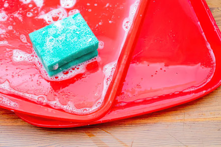 A green sponge on a red tray with soap suds on a wooden surfaceの写真素材