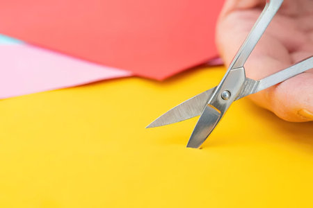 A hand holding scissors is cutting a yellow piece of paper with pink and red papers in the...