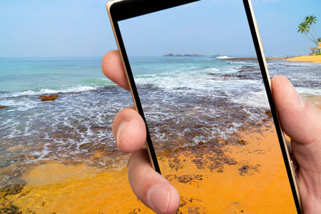 Hand holding smartphone to capture beautiful beach scene with calm blue ocean and sandy shore with...