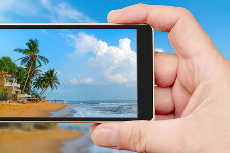 Hands holding a smartphone displaying a beautiful tropical beach with palm trees and a blue sky,...