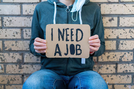 Man wearing a hoodie and jeans sitting against a brick wall holding a cardboard sign with the message I need a job.の写真素材