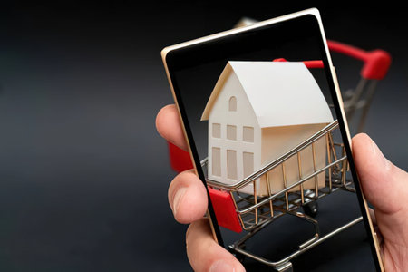 Person holds a smartphone showing a miniature house in a shopping cart representing online real...