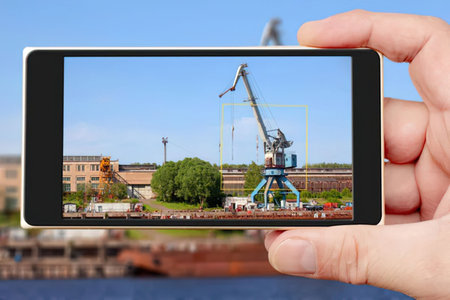 Person holds smartphone displaying augmented reality view of industrial crane in a dockyard with a...