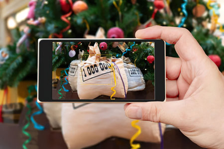 Person takes picture of christmas money gift bags under decorated tree with smartphone camera,...