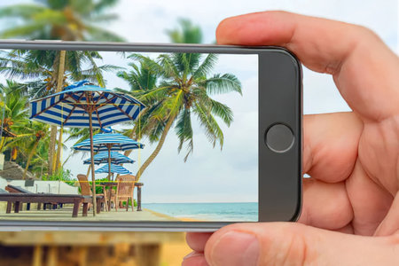 Hands holding smartphone displaying a serene tropical beach scene with palm trees and striped...