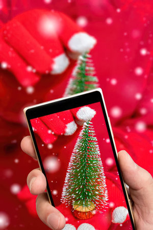 Hand holding smartphone showing miniature christmas tree on red background with snowfall symbolizing...