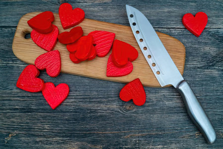 Red heart shaped objects are scattered on a wooden board with a sharp kitchen knife placed nearby on...