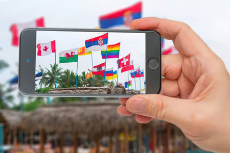 Person captures image of multiple international flags flying on a tropical area with blurry thatched...