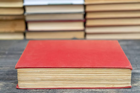 Red hardcover book lies on wooden table with stack of other books in the blurred background.
