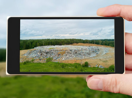 Hand holding smartphone displaying a rubbish dump with a forest in the background on a cloudy day symbolizing environmental concerns and pollution issues.の写真素材