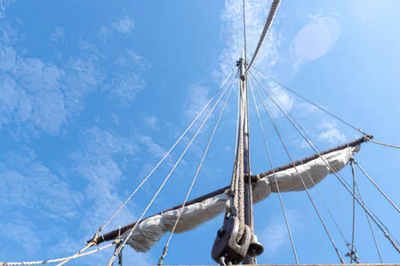 Majestic sailing ship mast with ropes and sail set against a vibrant blue sky with scattered clouds.の写真素材