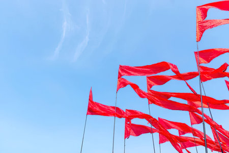 Multiple red flags are waving in the wind against a clear blue sky on tall poles.