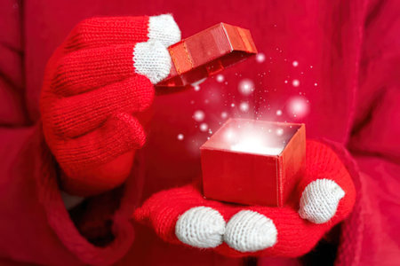 Person wearing red winter gloves holds a red gift box with sparkling contents inside, symbolizing a...