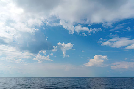 Calm ocean water reflects a bright blue sky with white fluffy clouds creating a serene and peaceful...