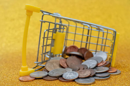 Miniature shopping cart spilling coins onto a textured golden surface, symbolizing retail...
