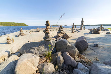 Numerous stone cairns are carefully balanced on a serene rocky shoreline with calm water and clear...