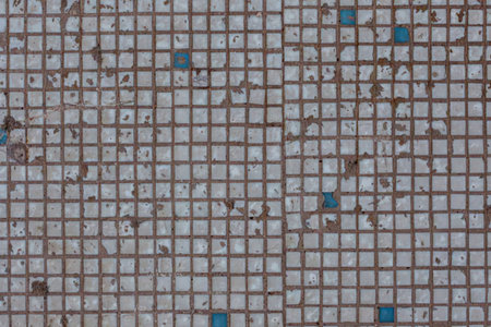 Worn white mosaic tiles with scattered blue accents on a grid pattern show signs of aging with dirty...