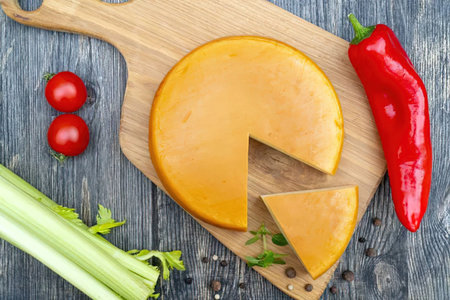 Orange cheese on a wooden board accompanied by fresh vegetables and spices on a rustic table,...