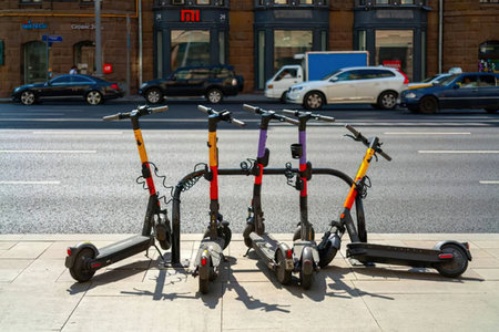 Five electric scooters are parked in a row on a city sidewalk alongside a busy street.